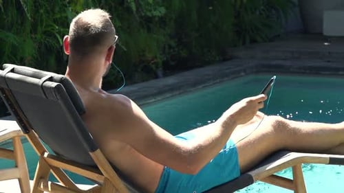 Young man enjoying music on sunbed by pool in summer super slow motion