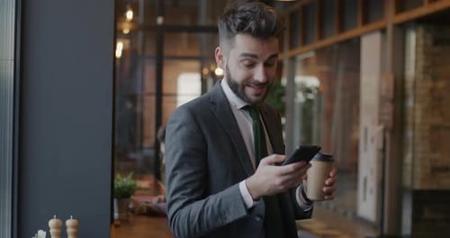 Slow Motion Portrait of Young Businessman Standing in Cafe Using Smartphone Holding to Go Coffee