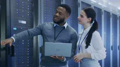 Black IT Technician with a Laptop Computer Gives a Tour to a Young Intern. They Talk in Data Center