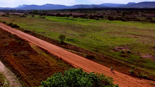 Drone, solo cyclist rides isolated dirt road through rural farming landscape