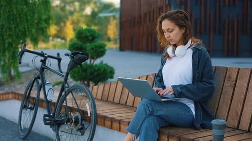 Kaukasisches lächelndes Mädchen sitzt auf der Bank und arbeitet am Laptop in der Nähe ihres Sportfahrrads