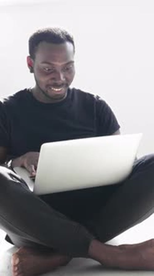 Homme afro-américain assis les jambes croisées utilisant un ordinateur portable dans un espace intérieur minimaliste lumineux