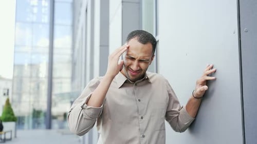 A young adult man feels a headache while standing on the street near an office building. Sad mixed