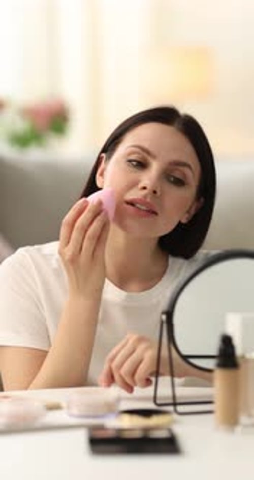 Young woman applying foundation with makeup sponge near mirror at table indoors