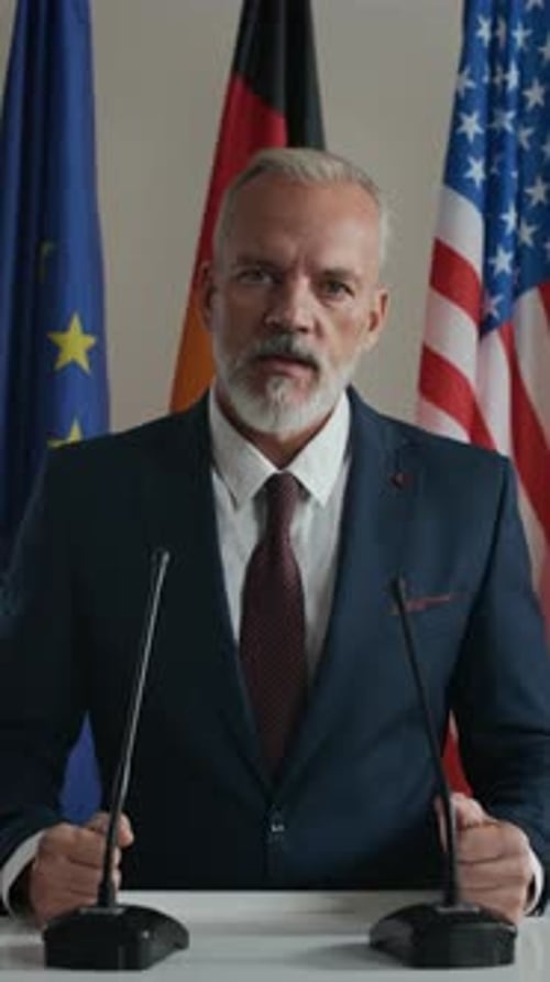 Man in Suit Speaking Behind Microphones with Flags