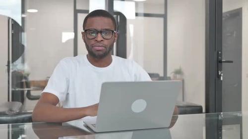 Young African Man Looking at Camera while Working on Laptop in Office