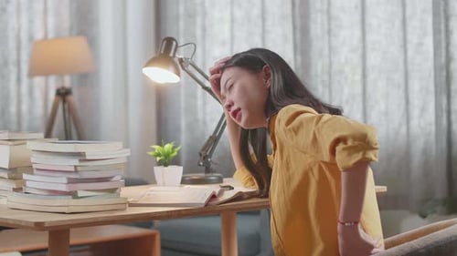 Side View Of Young Asian Female Studying At Home Having A Backache While Reading Books On The Table