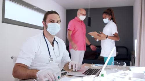 Doctor Working on Laptop With Patient in Background