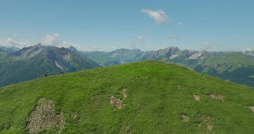 Two hikers trek a grassy hilltop with a vast mountain range in the background under a clear sky