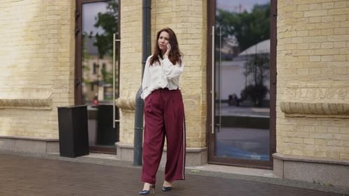 A Stylishly Dressed Businesswoman Walks and Talks on a Mobile Phone Against the Backdrop of a City