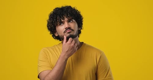 Pensive Young Indian Man Thinking with Hand Near Chin Standing Over Orange Studio Background