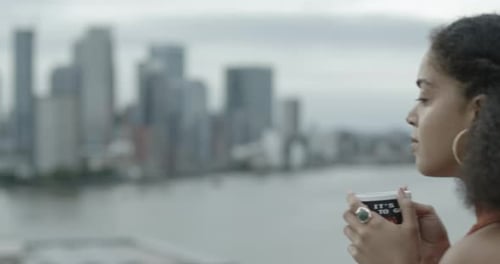 Pretty young woman on balcony looking out over city at sunset drinking a cup of tea