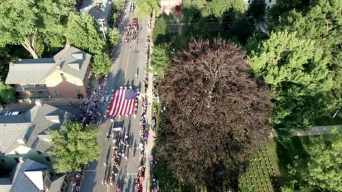 Aerial of crowds lining streets of small town parade, US flags fill grassy lawn, flag carried by Boy