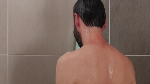 A STATIC CLOSE UP SHOT of a man standing, having a relaxing shower and running his hands through his