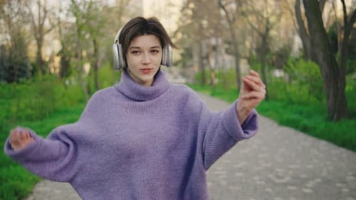 Woman Dances on a Paved Path in a Park While Wearing Headphones The Sun Shines Down and Trees