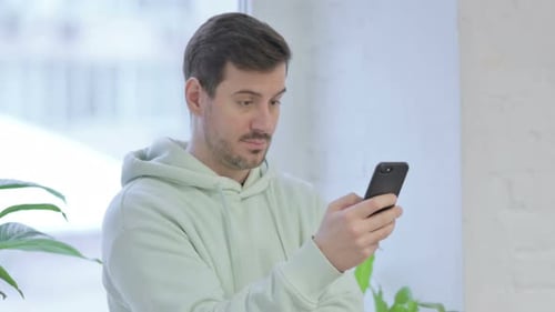 Young Man Using Smartphone while Standing Near Window