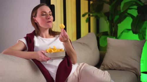 Young Woman is Relaxing on the Couch Watching TV and Eating Chips