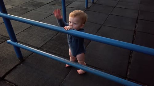 Happy toddler boy stands alone on rubber playground mat leaning against blue jungle gym looking over