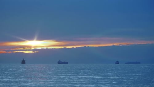 Steady sunset shot with a flare of a three cargo ships sailing in a still water near port