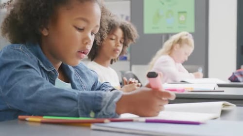 Video of biracial schoolgirl sitting at desk writing in diverse school class