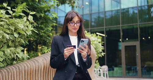 Young Woman Using Phone and Credit Card for Online Banking While Sitting Near a Glass Office