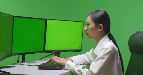 Side View Of Asian Woman Working With Mock Up Multiple Computer Monitor In Green Screen Studio