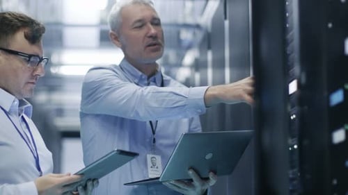 Two IT Technicians Standing in Working Data Center. They Use Laptop and Tablet Computer while Stand