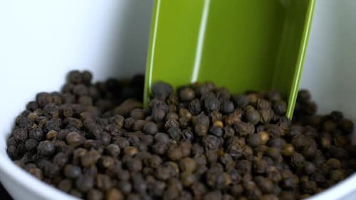 Close-up of whole black peppercorns inside of a white bowl. A green plastic spoon stirs the spice.