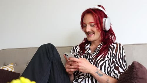 Non-binary person wearing headphones using smartphone on couch