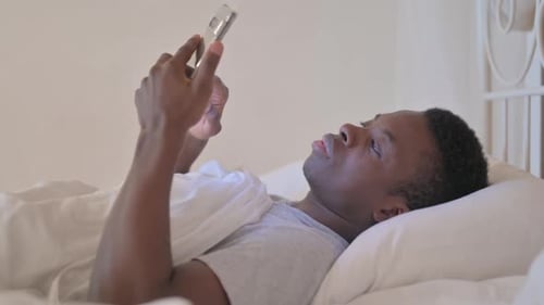 Side View of Young African Man using Phone in Bed
