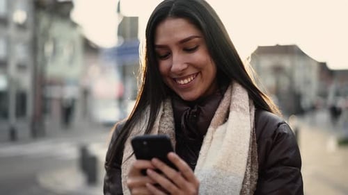 Happy woman holding phone outside in city street. Smiling young female adult girl liking text