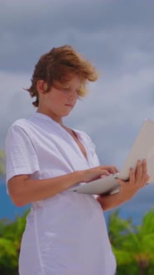 Vidéo verticale Jeune garçon debout sur la plage avec un ordinateur portable faisant ses devoirs ou parlant à quelqu'un