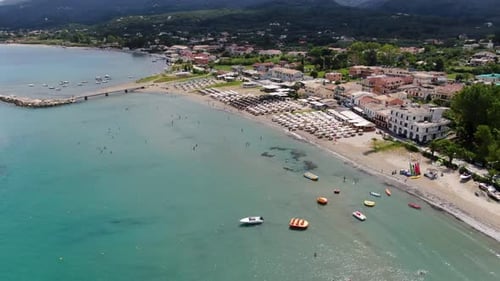 Lively aerial view of a bustling beach and coastal town in Corfu, Greece, with colorful boats,