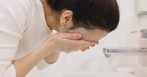 Mujer lavándose la mascarilla con agua en un baño blanco