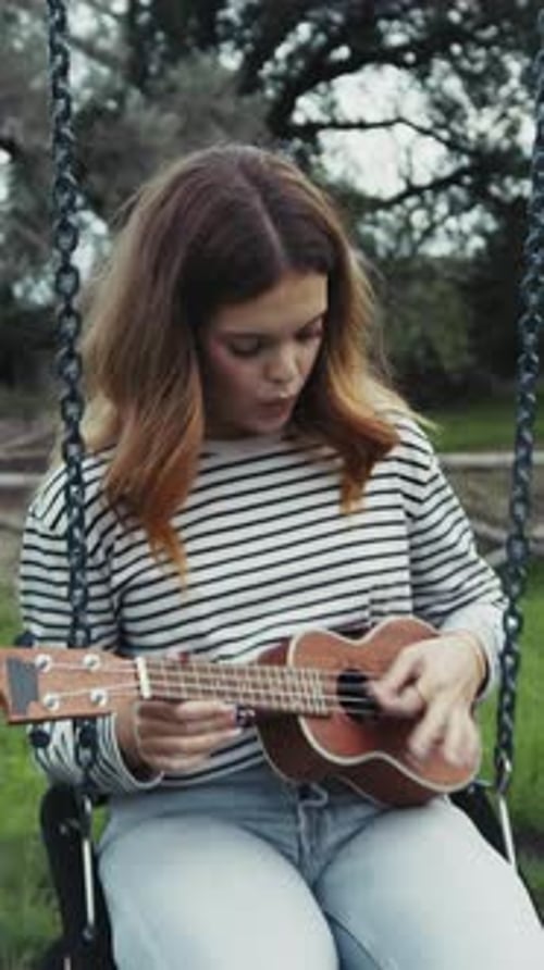 Young Woman Practices Playing The Ukulele Seated In A Swing