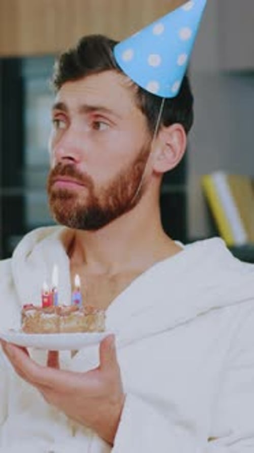 Unhappy Sad Young Man Celebrates Birthday Alone Hold Cake Watch TV Sitting at Sofa in the Living
