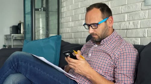 Young Man Calculating Bills on Smartphone Sitting on Sofa 30s