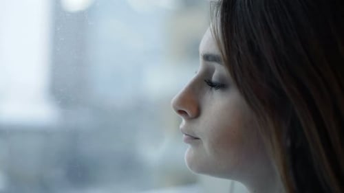 Pensive young woman looking out window on overcast day