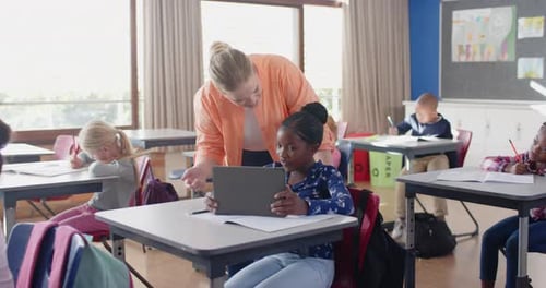 In school, female teacher helping student with tablet in classroom, other students studying