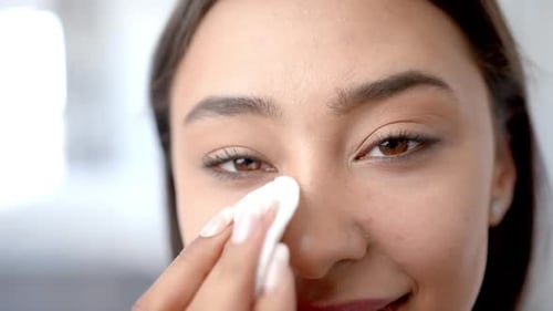 Smiling woman cleaning face with cotton pad, focusing on skincare routine