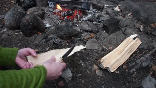 White male whittle chips from wood log with finnish puukko knife. Green shirt. Camp fire on the back