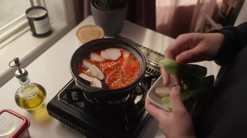 Adding Vegetables to Simmering Red Soup in Clay Pot on Portable Gas Stove