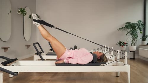 Woman In Workout Clothes Performs Leg Exercises On Pilates Reformer At Gym. wide static shot