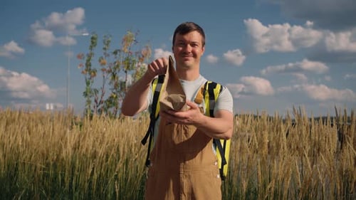 Un repartidor sonriente con uniforme y mochila ofrece una bolsa de papel a la cámara en un parque soleado junto a un dorado