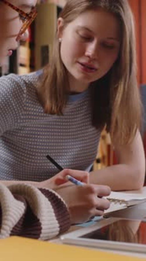 Two Female Group Mates Discussing Assignment in Workbook in College Library