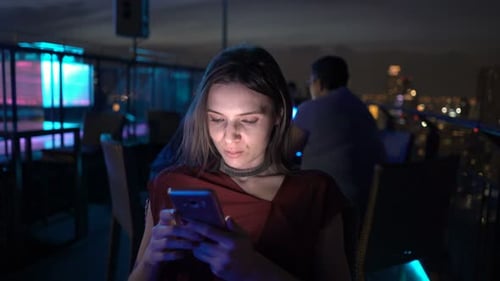 Young woman texting on smartphone on rooftop terrace at night in the city