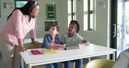 African American teacher walking left, leaning and pointing at tablet, coaching two teenage girls