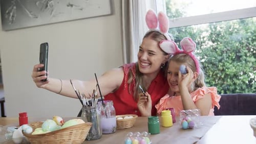 Mom and daughter taking selfie with mom extending phone while painting eggs at table for Easter