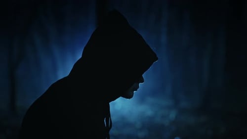 A Dark Figure in a Cloak with a Hood in a Sinister Night Forest Fantasy Demon or Witcher in the
