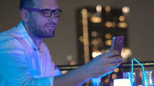 Young man enjoys cocktail and takes selfie on rooftop terrace at night